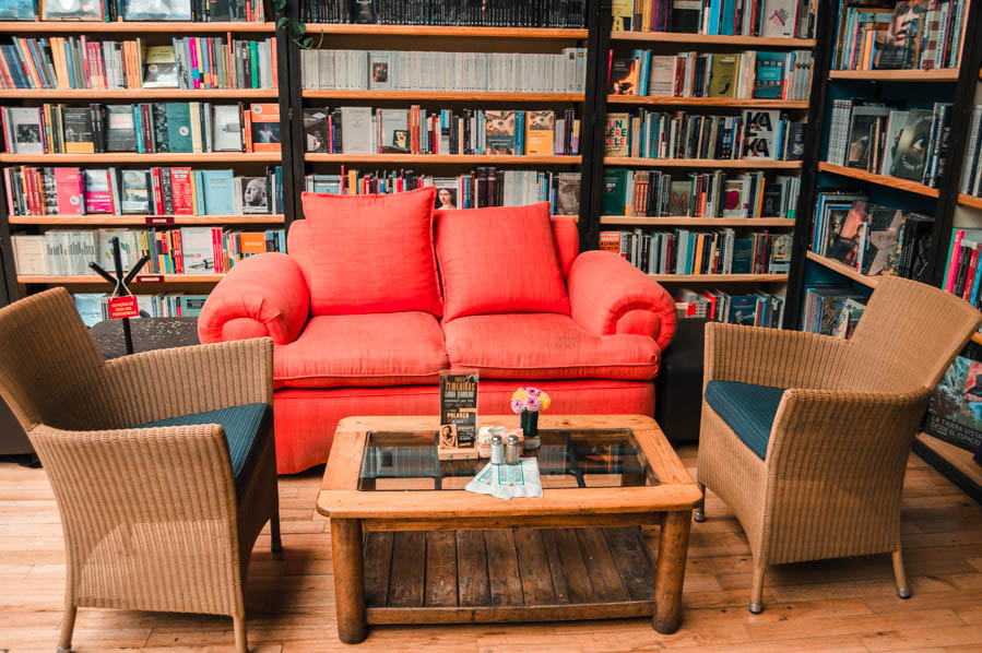 A comfortable reading nook in Cafebrería El Péndulo, featuring a bright red sofa, wicker chairs, and a wooden coffee table surrounded by bookshelves.