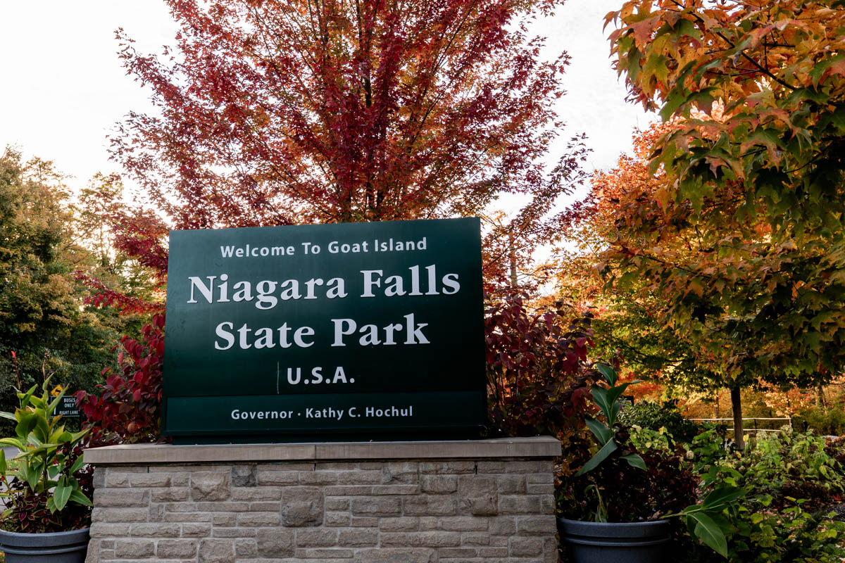 Entrance sign to Niagara Falls State Park on Goat Island, surrounded by vibrant red and orange fall foliage.