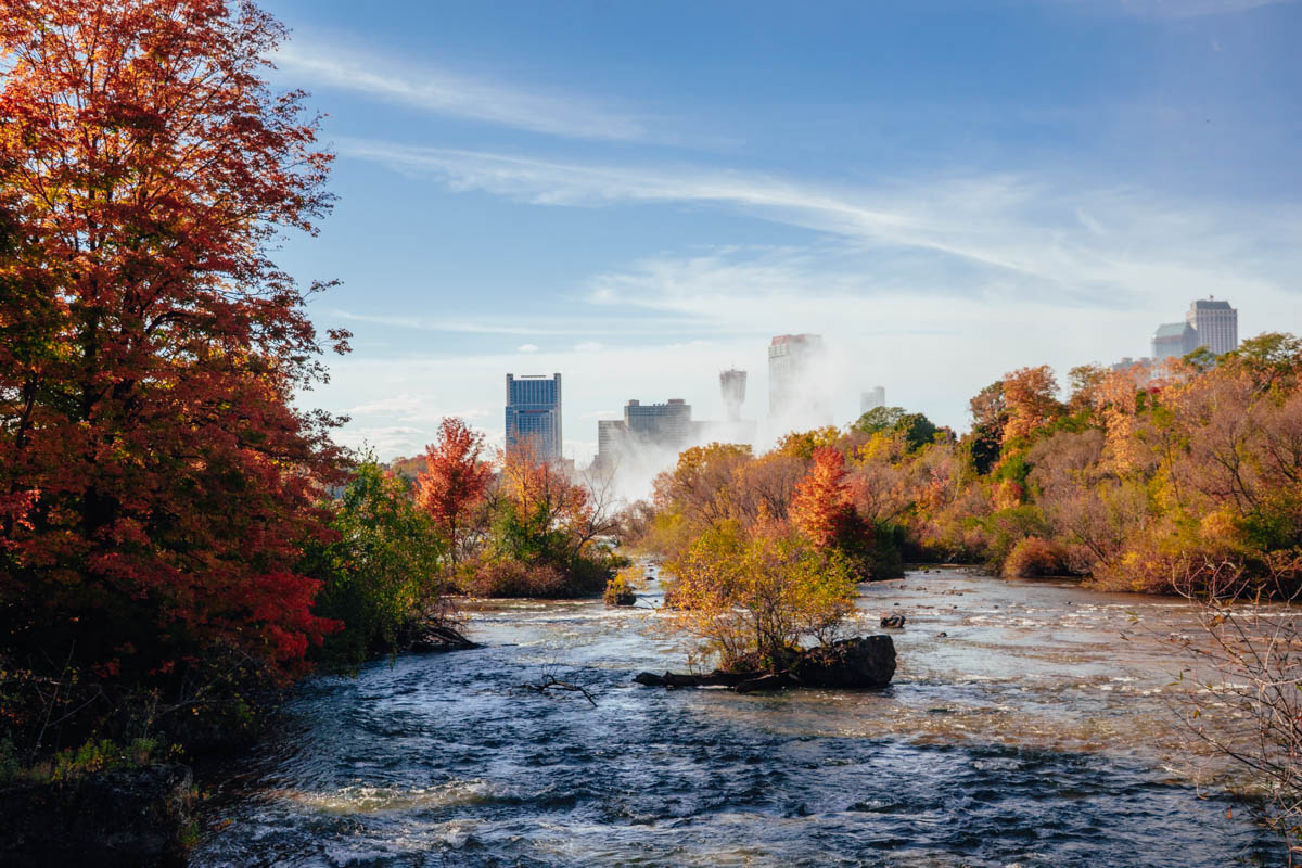 View of Niagara Falls skyline through vibrant autumn trees along a peaceful river in Niagara Falls State Park.