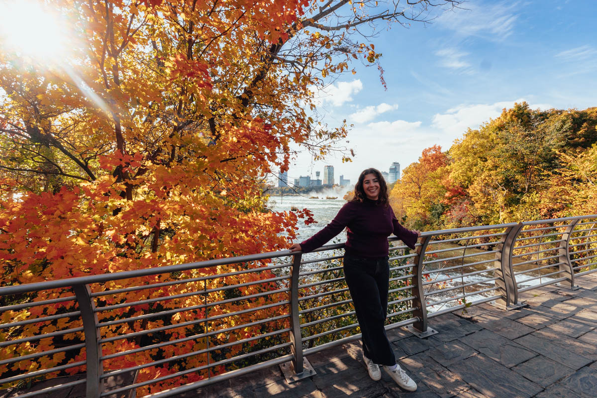 Woman smiles beneath a canopy of golden fall leaves at Niagara Falls, with the skyline and river in the background.