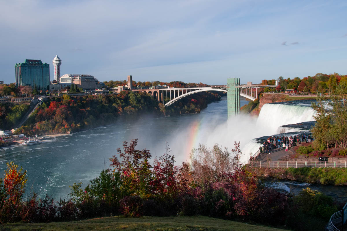Panoramic view of Niagara Falls with a vibrant rainbow arcing above the American Falls, surrounded by fall foliage and the Canadian skyline in the background.