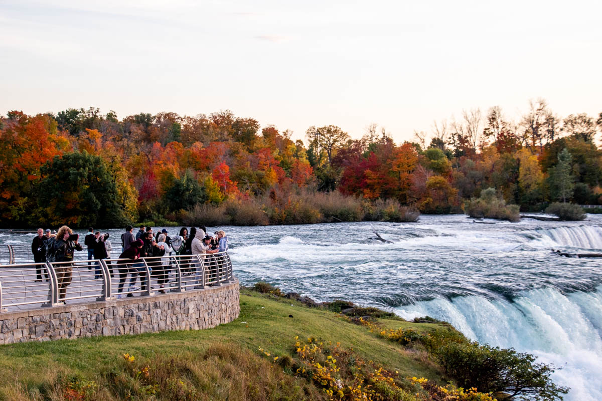 Tourists gather at a scenic overlook near the rapids, with colorful fall trees lining the river in the background.
