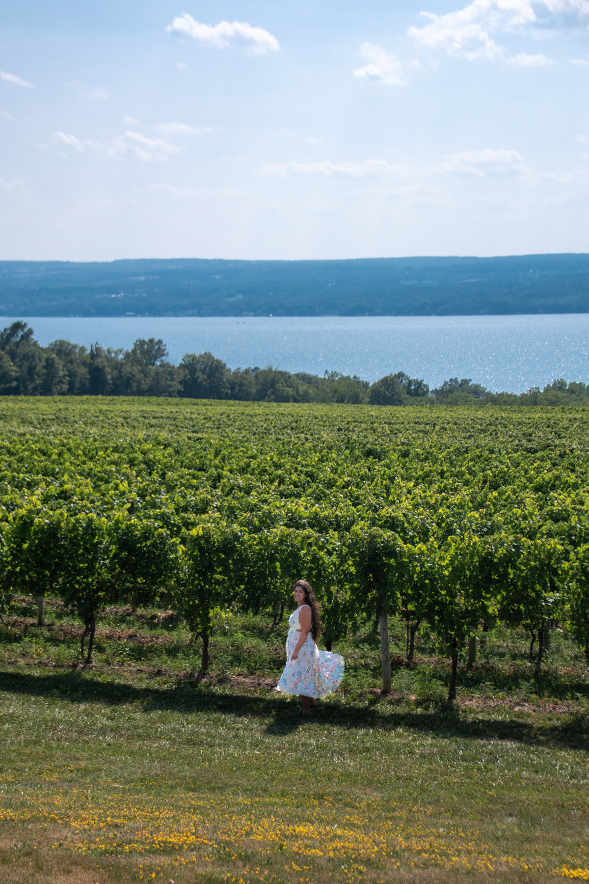 Woman in a floral dress walking through vineyards with a view of Seneca Lake in the distance.