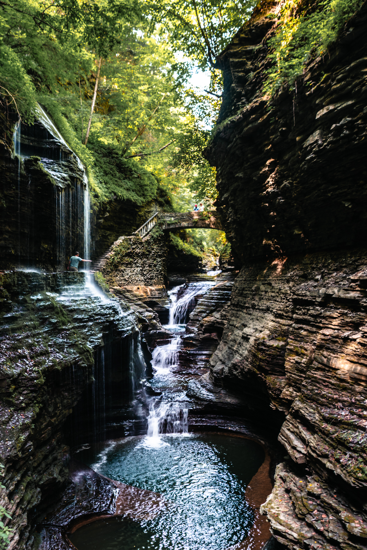 Multiple waterfalls flowing into emerald pools beneath a stone bridge in the gorge at Watkins Glen State Park.