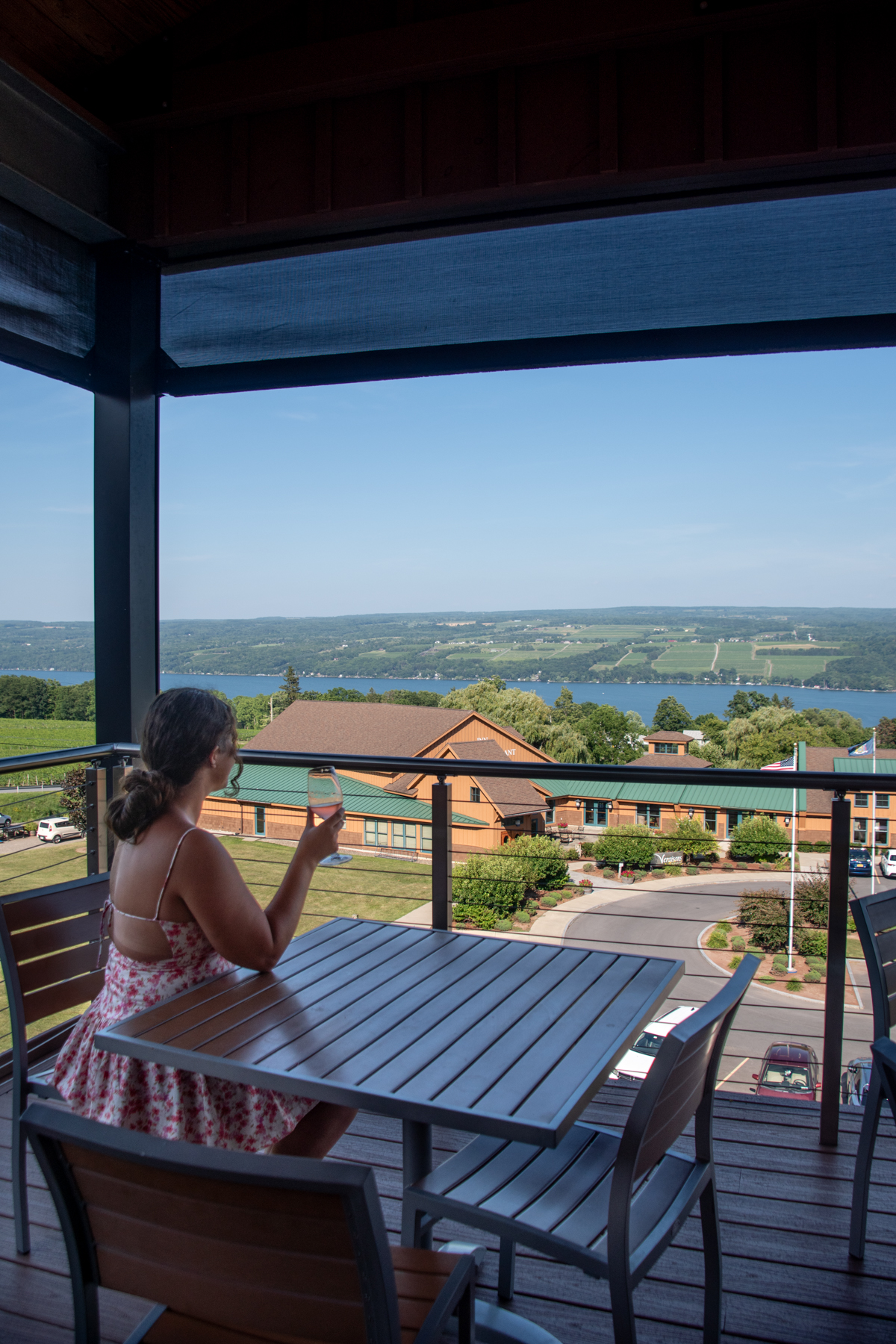 Woman in a floral dress enjoying a glass of wine on an outdoor balcony with sweeping views of Seneca Lake and surrounding vineyards.
