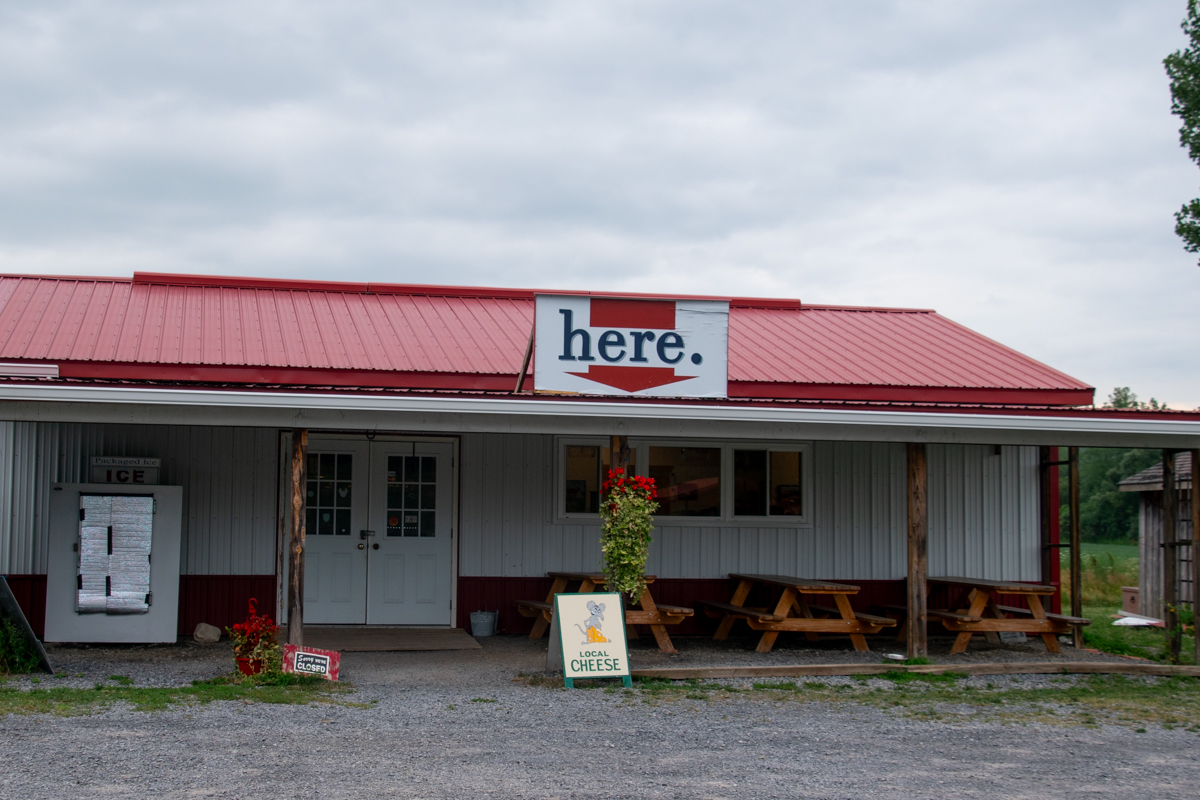 Exterior of a market-style building with a red metal roof, “here.” sign, and picnic tables out front.
