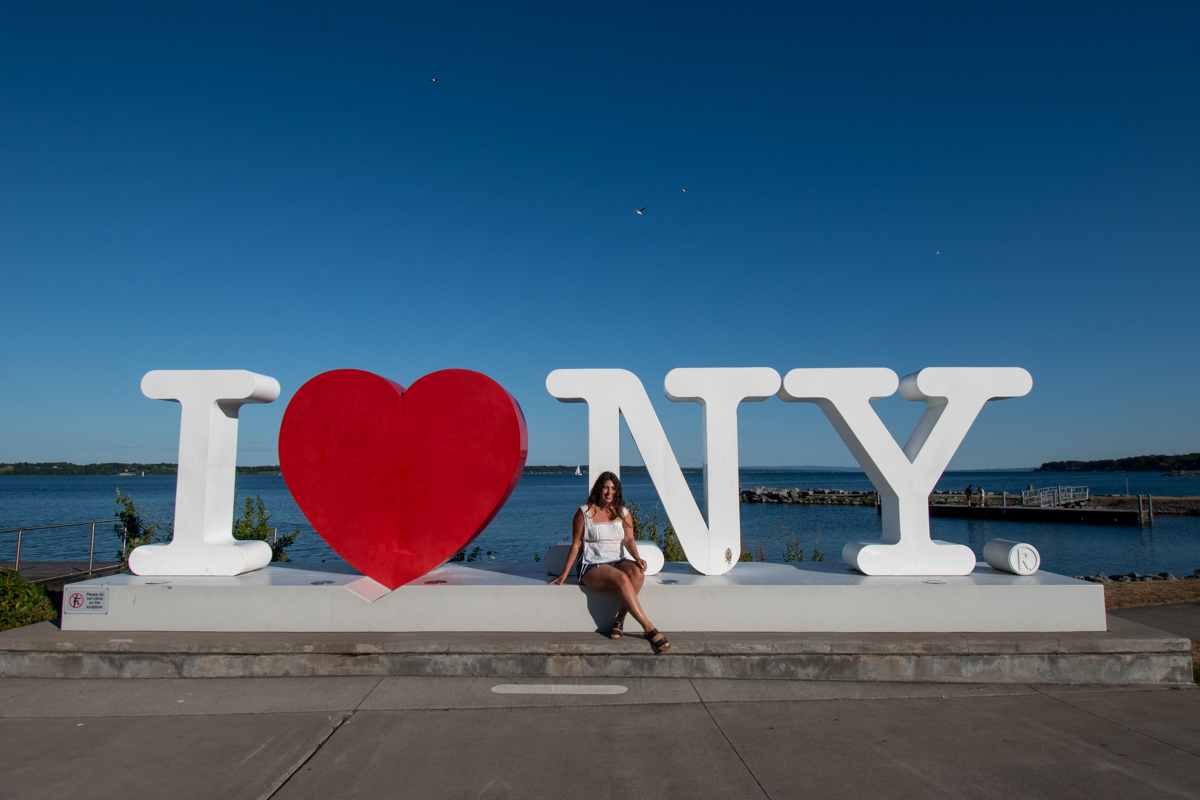 Woman sitting on the “I ❤️ NY” sign by the Seneca Lake waterfront in Geneva, New York, on a sunny day.