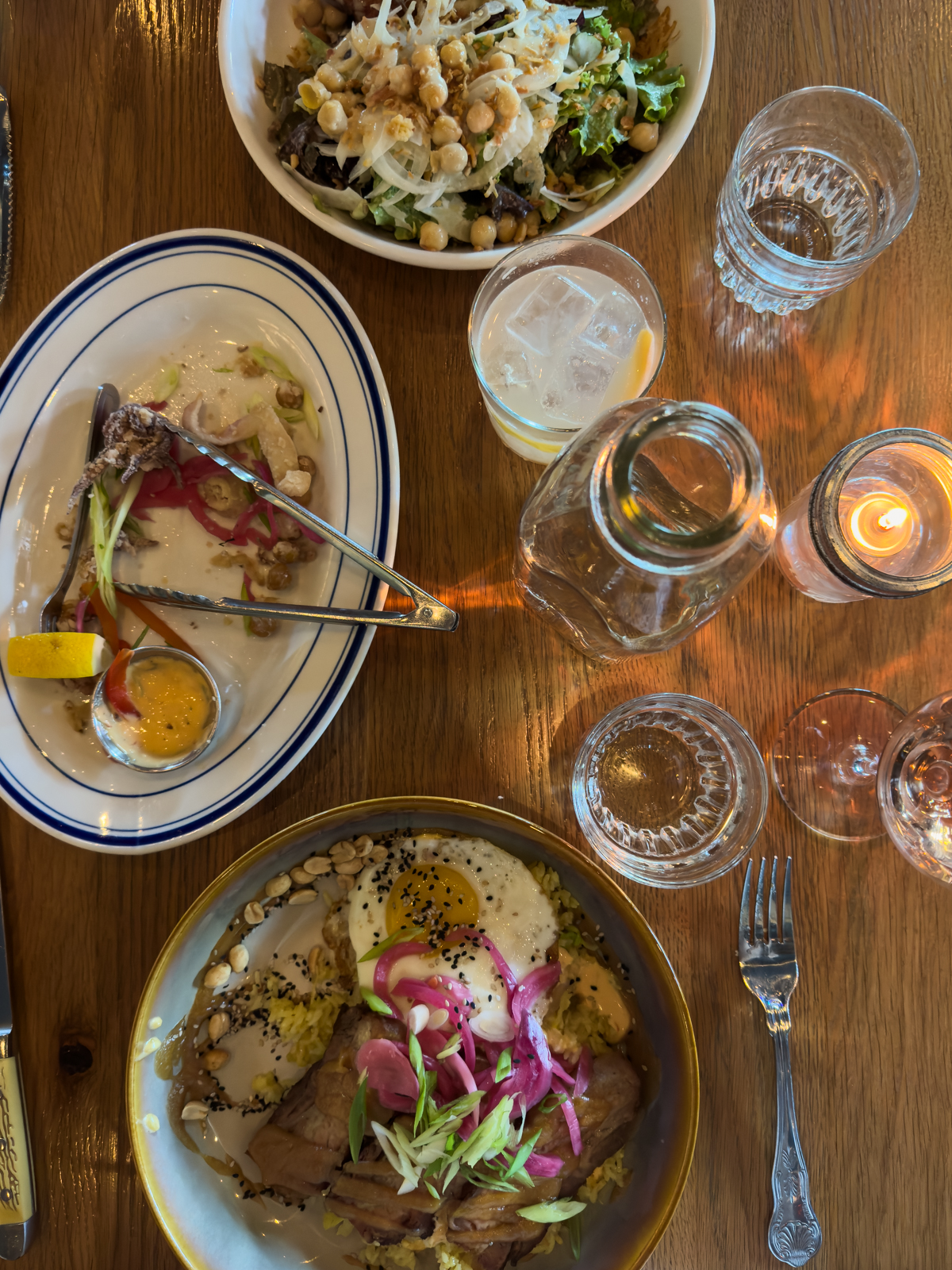 Overhead view of a colorful meal with a salad, grain bowl topped with egg, pickled onions, and grilled meat, alongside drinks and candles on a wooden table.