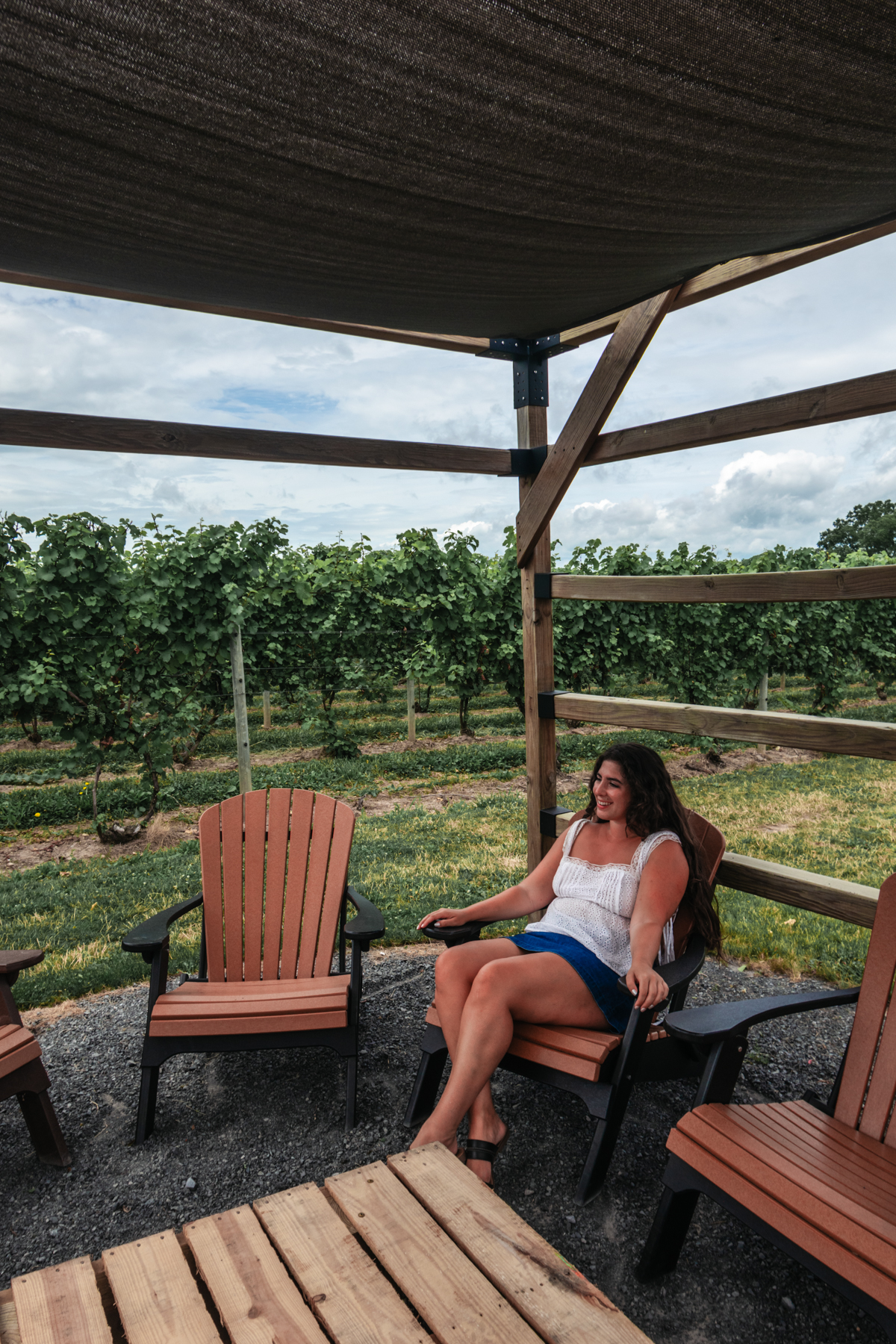 Woman relaxing in an Adirondack chair under a shaded wooden pergola with vineyard rows in the background at a Finger Lakes winery.