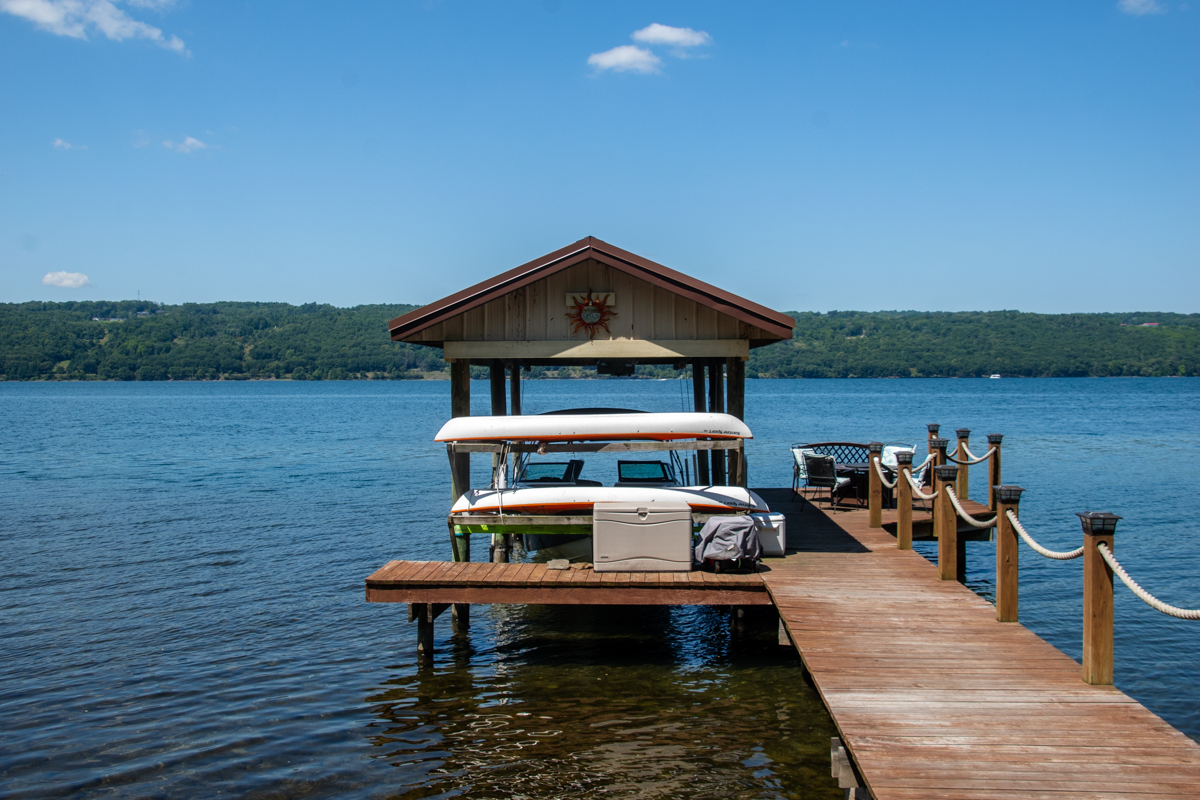 Boat dock on Seneca Lake – A wooden dock with rope railings extends over Seneca Lake, leading to a covered boat slip with a motorboat beneath, surrounded by calm water and distant green hills.