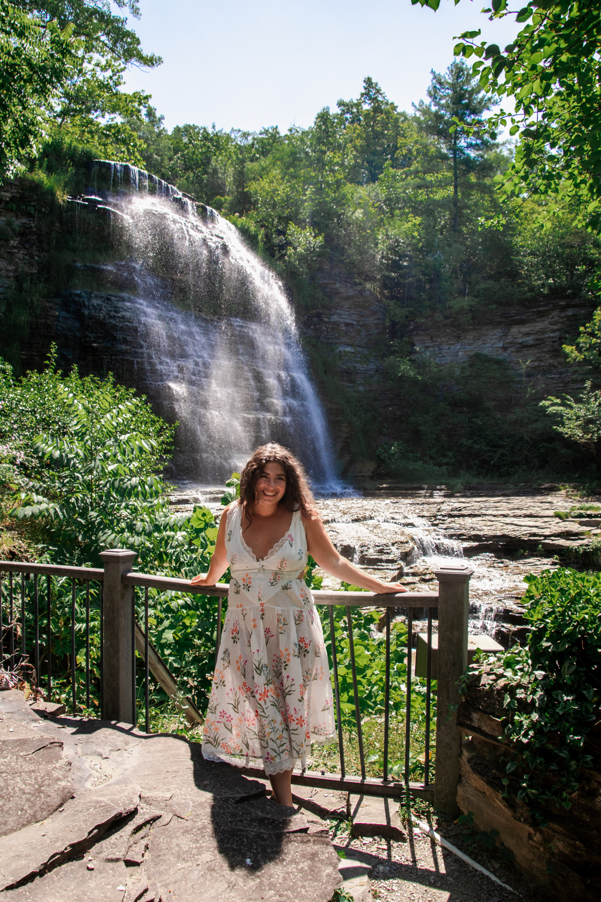 Woman in a white floral dress smiling in front of a cascading waterfall surrounded by lush green trees at a scenic overlook.