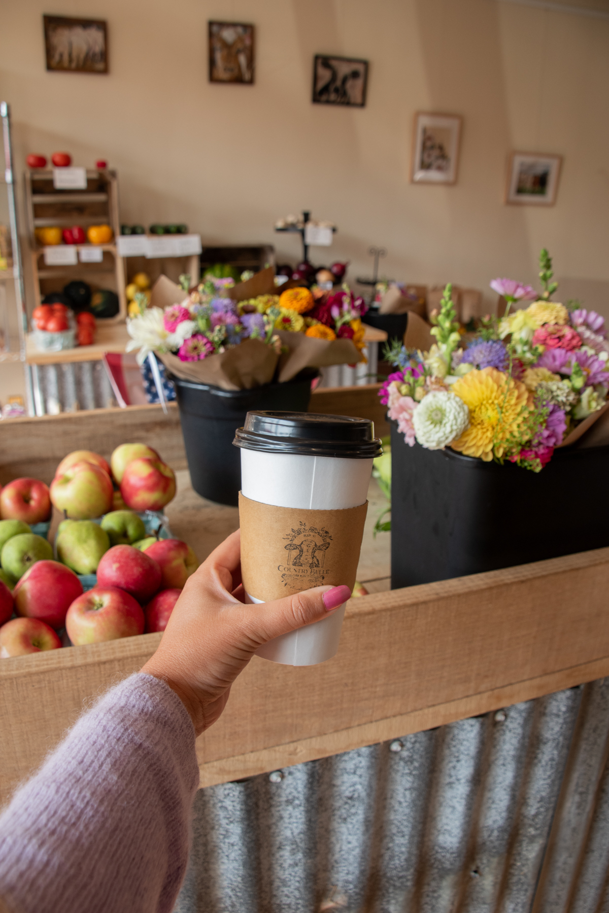 Coffee and flowers at Country Belle Farm Market – A hand holding a to-go latte in front of colorful bouquets and baskets of fresh apples inside the farm market, showcasing local produce and fresh blooms.