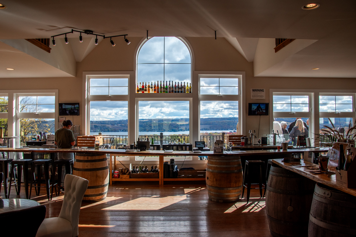 A bright tasting room at Vineyard View Winery with large arched windows overlooking Keuka Lake and fall foliage on the Keuka Lake Wine Trail.