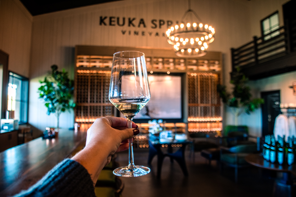 A hand raises a glass of white wine inside Keuka Spring Vineyards, capturing the warm glow of the tasting room lighting and the stylish wine wall behind it.