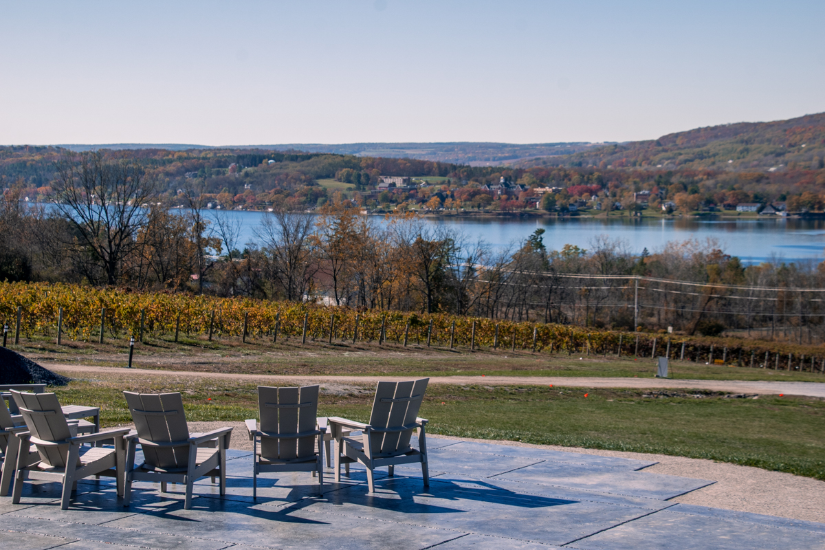Adirondack chairs arranged on an outdoor patio at Keuka Spring Vineyards, overlooking vineyard rows and the blue waters of Keuka Lake under a clear sky — a peaceful wine tasting setting.