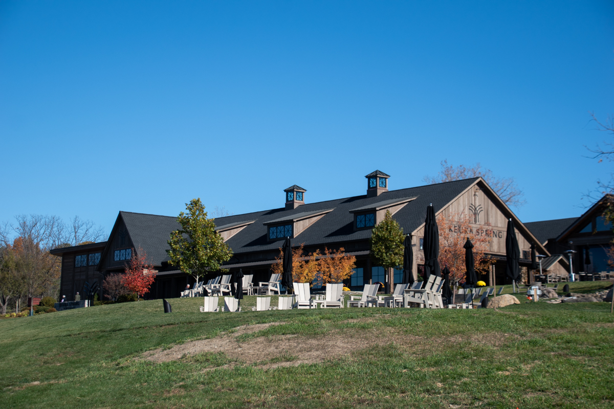 Wide view of Keuka Spring Vineyards’ large wooden winery building beneath a bright blue sky, with outdoor seating and autumn foliage in the foreground. A scenic Finger Lakes winery visit.