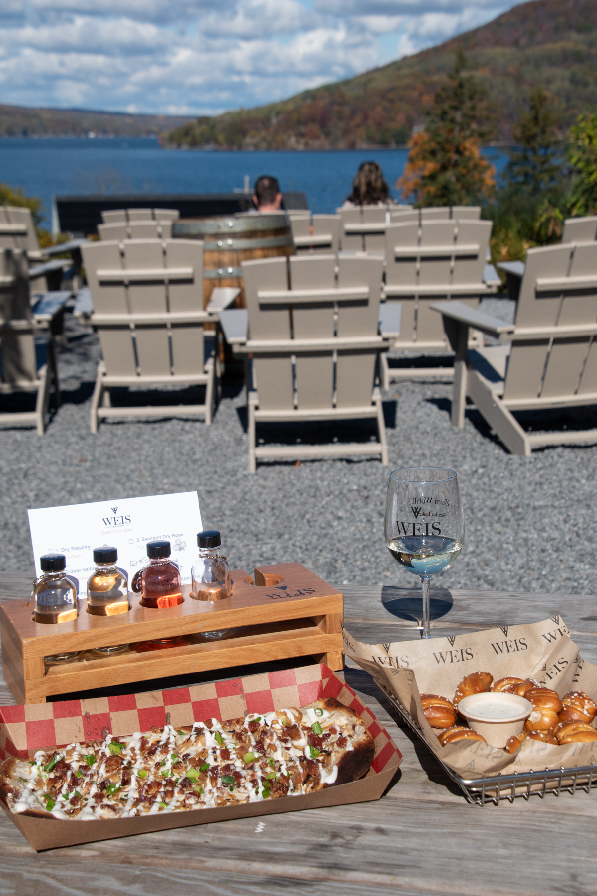 Wine flight, pretzel bites, and a Flammkuchen flatbread arranged on a picnic table at Weis Vineyards, with lake views in the background