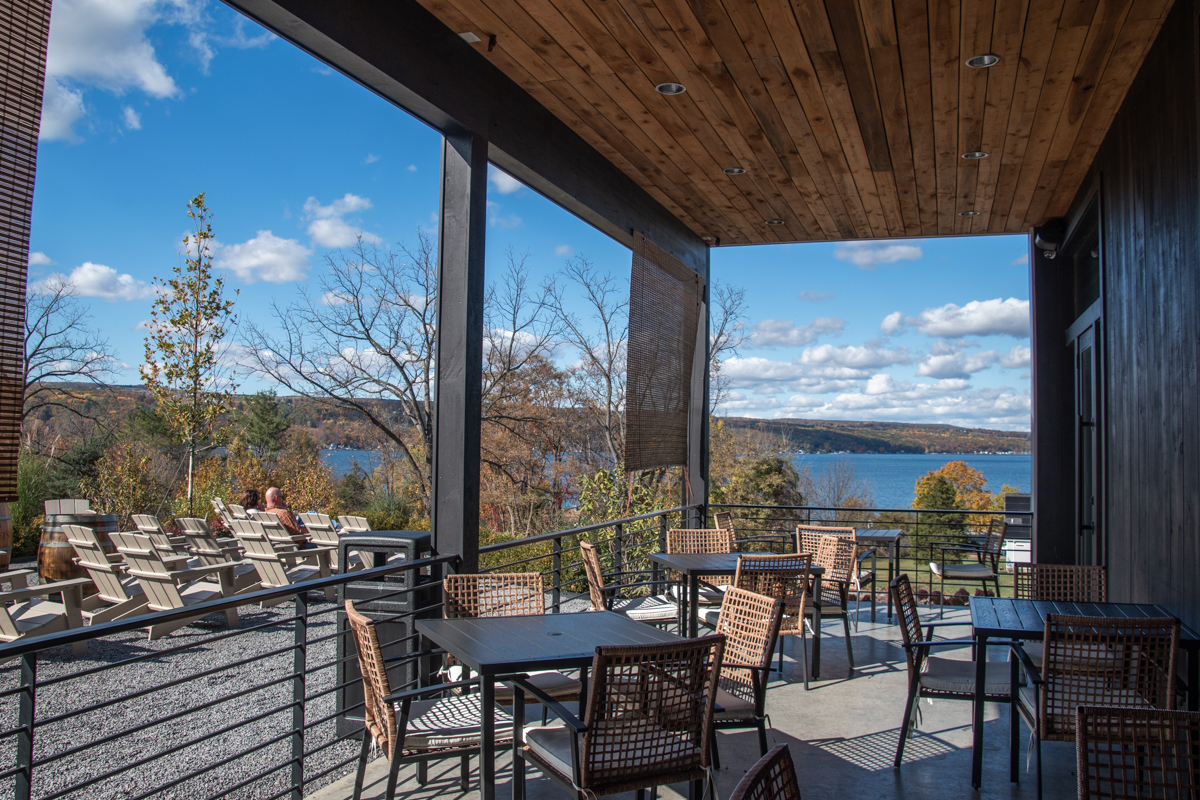 “Outdoor patio seating at Weis Vineyards overlooking Keuka Lake, with woven chairs, black tables, and Adirondack chairs arranged near the edge of the hill. Bright blue sky with scattered clouds and fall foliage frame the panoramic lake views along the Keuka Lake Wine Trail