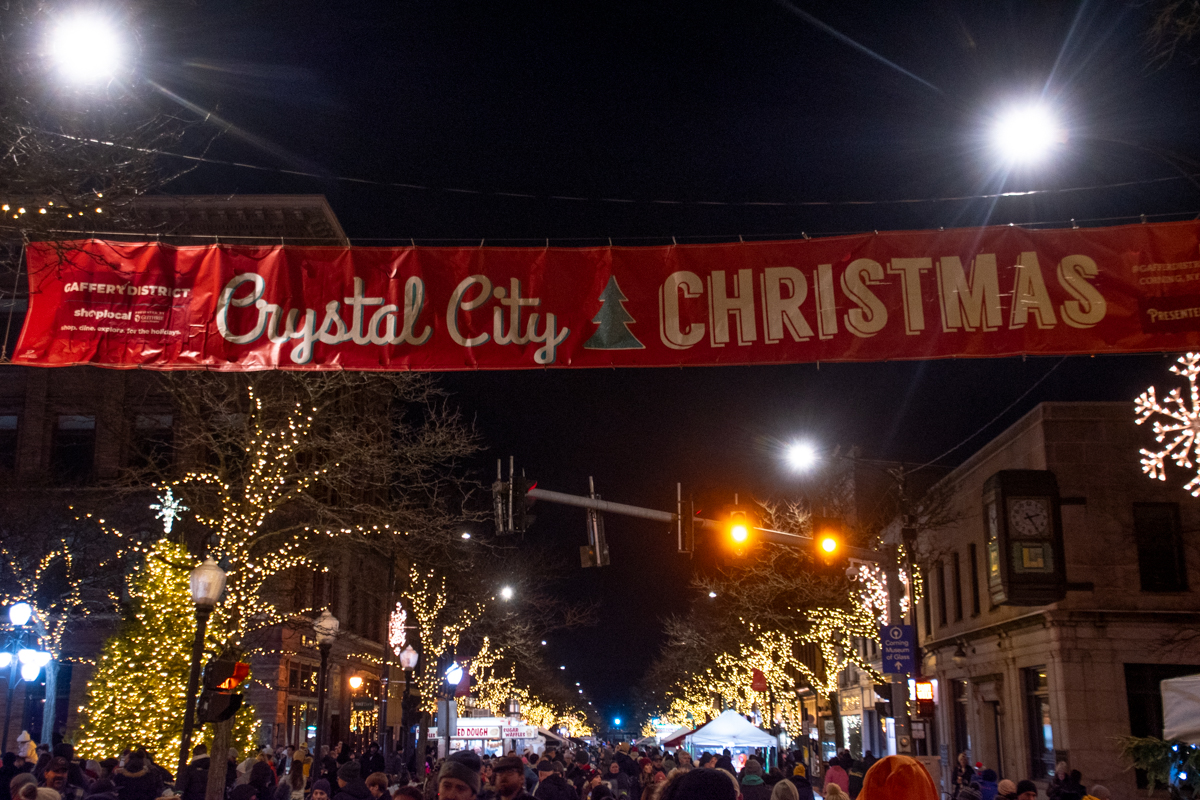 A red “Crystal City Christmas” banner hangs above a festive street in Corning, NY, where trees wrapped in sparkling lights and holiday market tents line the road as crowds gather at night.