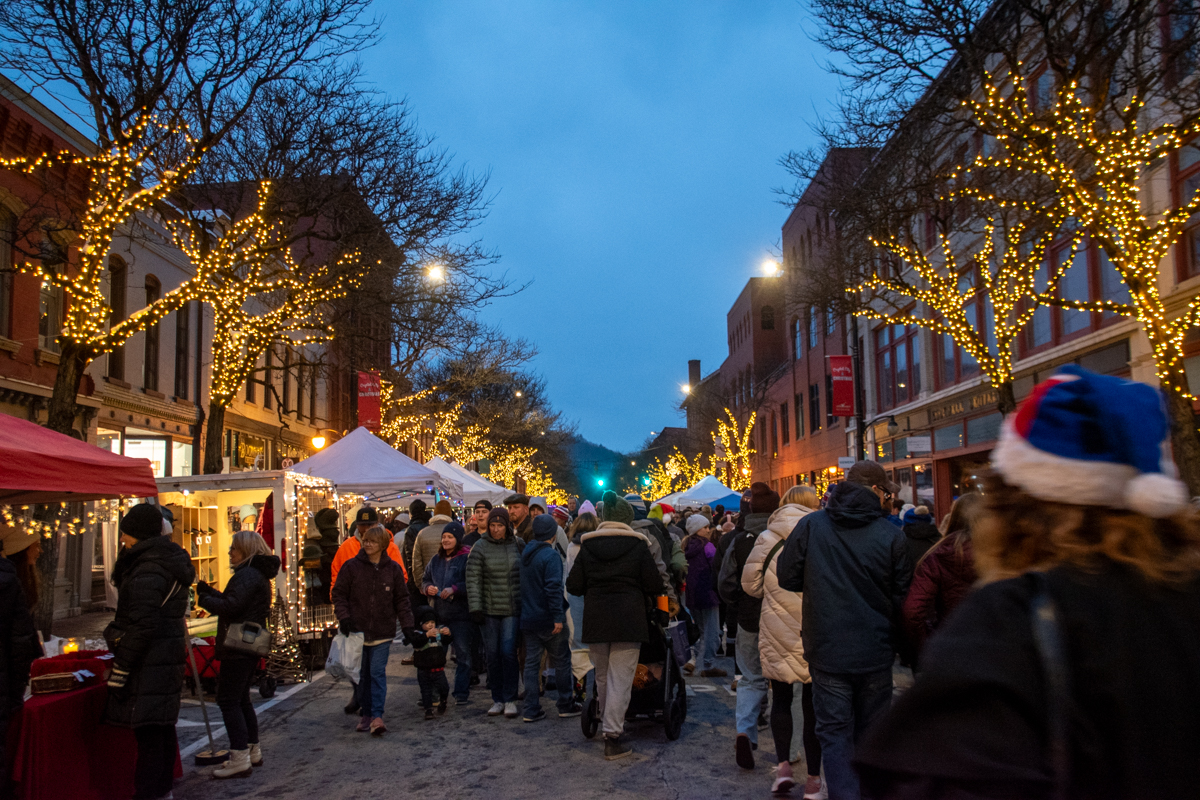 A busy winter street scene in downtown Corning, NY, with people in coats walking between white vendor tents and trees wrapped in golden Christmas lights at dusk.