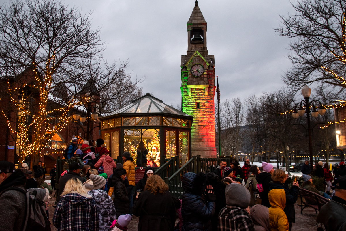 A crowd gathers in a festive square in Corning, NY, as families line up to see Santa inside a glass pavilion decorated with Christmas lights, with the illuminated historic clock tower glowing red and green in the background.
