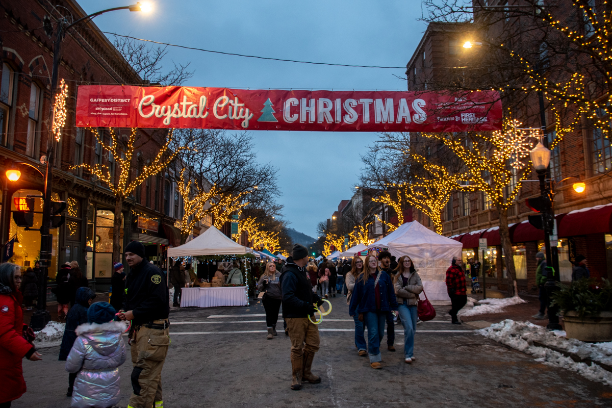 A festive nighttime scene on Market Street in Corning, NY, with the “Crystal City Christmas” banner stretched overhead and trees wrapped in twinkling lights illuminating the street.
