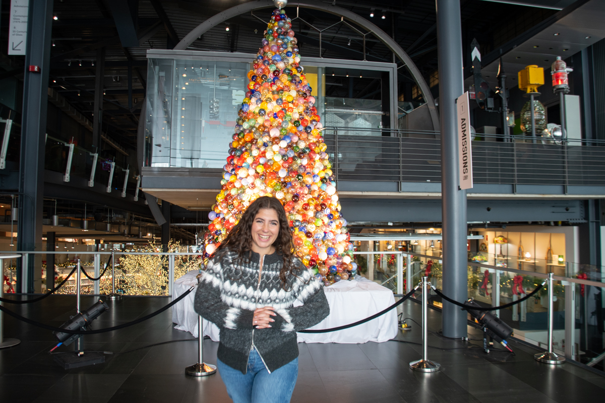 Woman smiling and posing in front of a large Christmas tree made of colorful blown-glass ornaments at the Corning Museum of Glass during Sparkle in Corning, NY.