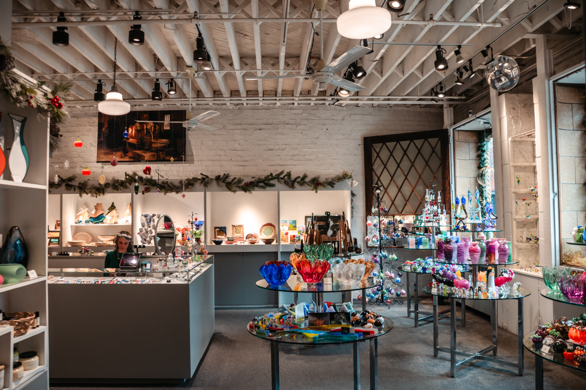Interior of Vitrix Hot Glass Studio with holiday décor, glass art displays, and an artist working behind the counter in Corning, NY.