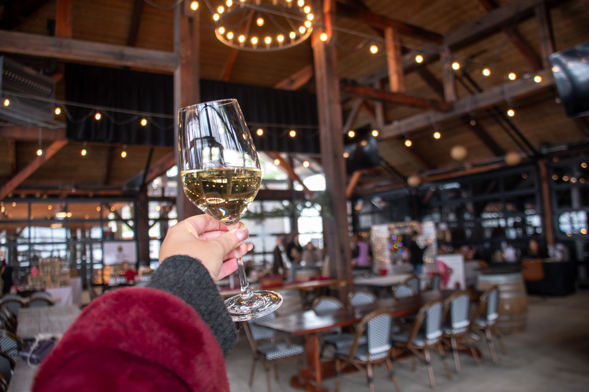Hand holding a glass of white wine inside the pavilion at Point of the Bluff Vineyards Holiday Market in Hammondsport, NY, with string lights and vendor tables in the background.