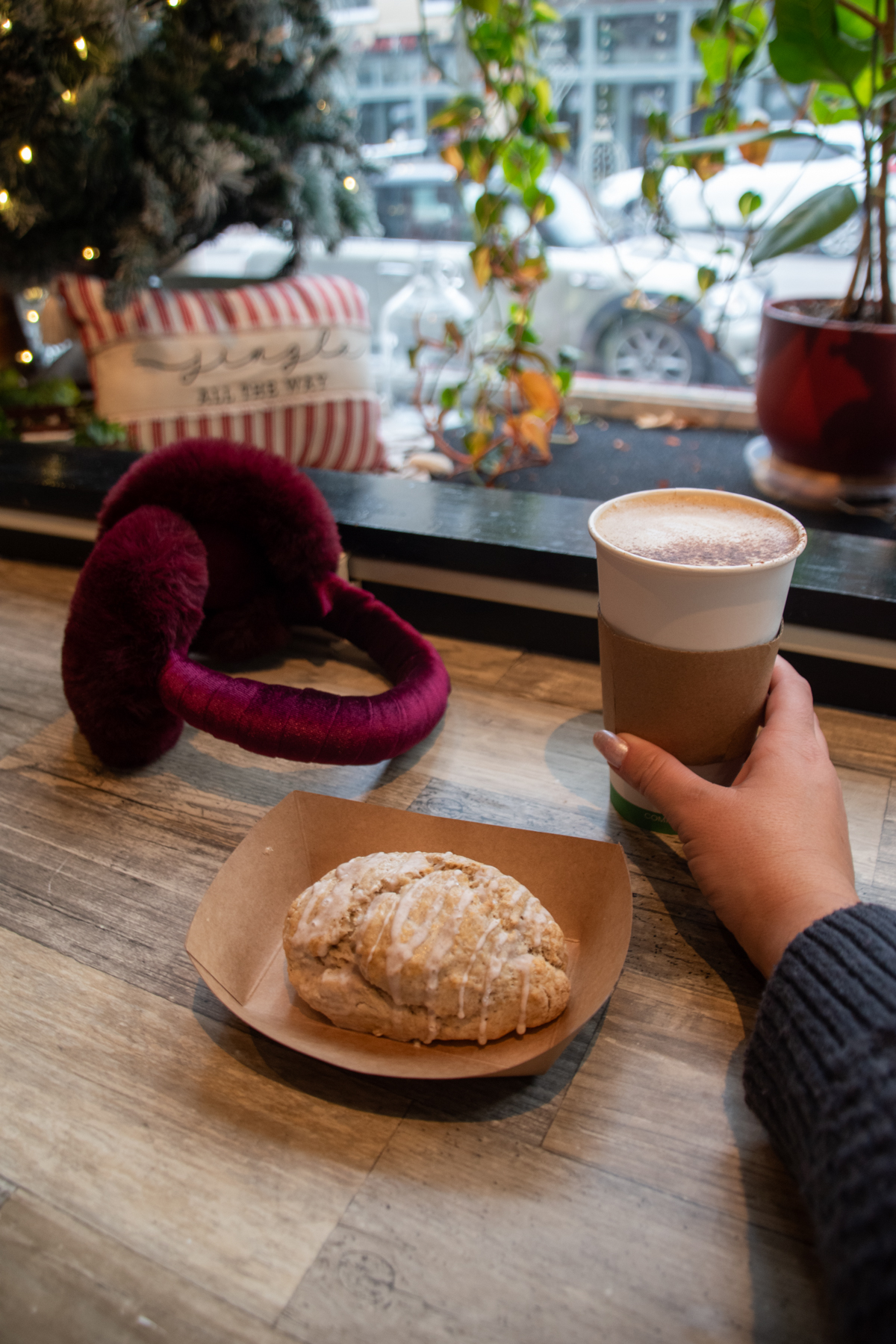 Hand holding a latte beside an eggnog scone at Homestead Bakery in Corning, NY, with winter décor, plants, and a snowy street view seen through the window.