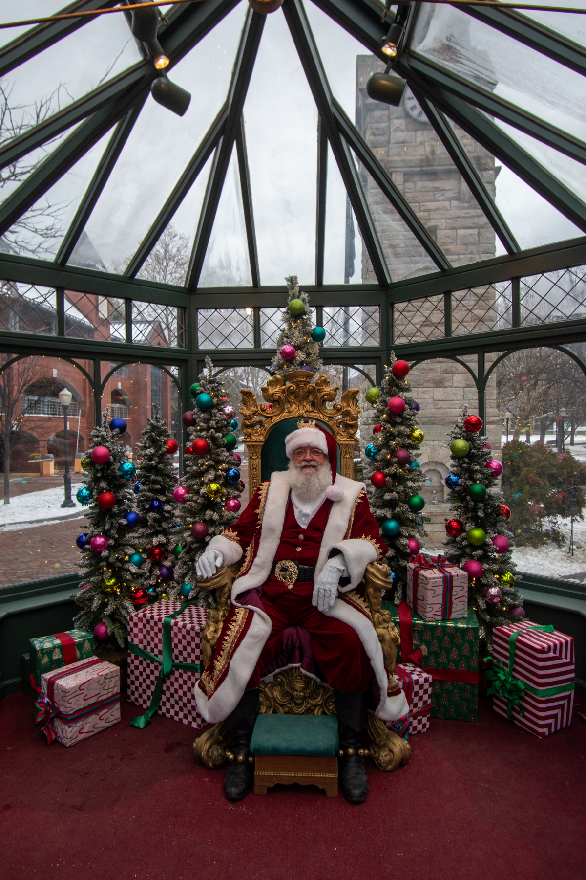 Santa Claus seated on an ornate golden chair inside a festive glass pavilion decorated with colorful ornaments, Christmas trees, and wrapped gifts in Corning, NY.