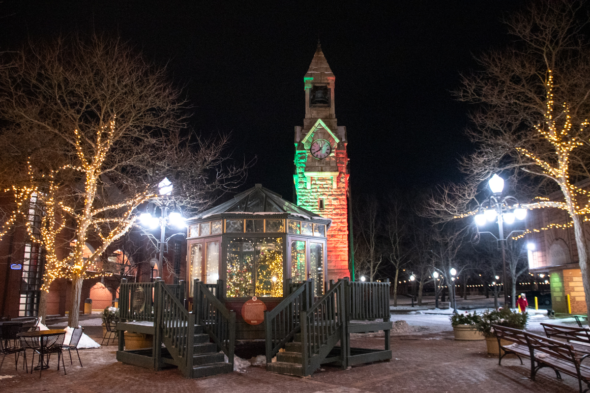 Illuminated glass pavilion decorated with Christmas lights in front of Corning’s Old City Hall clock tower at night, with trees wrapped in twinkling lights.