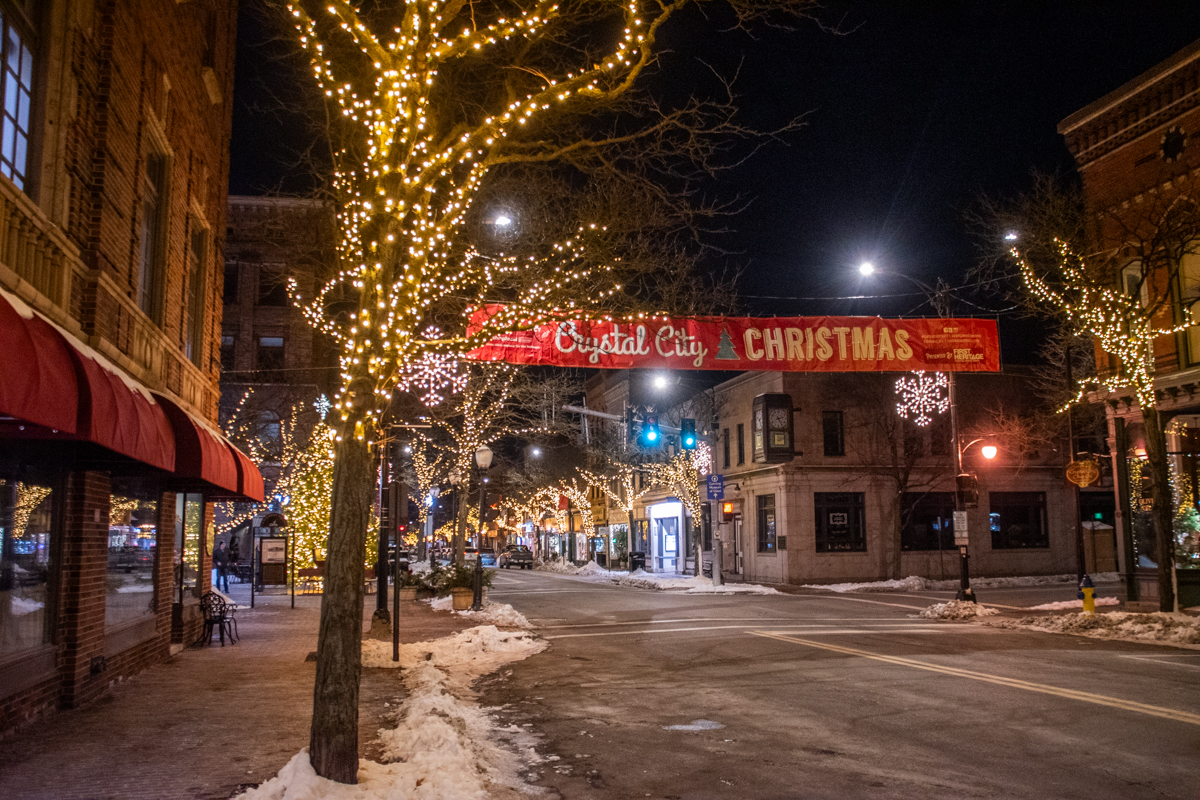 A nighttime street scene in Corning, NY with trees wrapped in warm white lights and a red “Crystal City Christmas” banner hanging over Market Street.