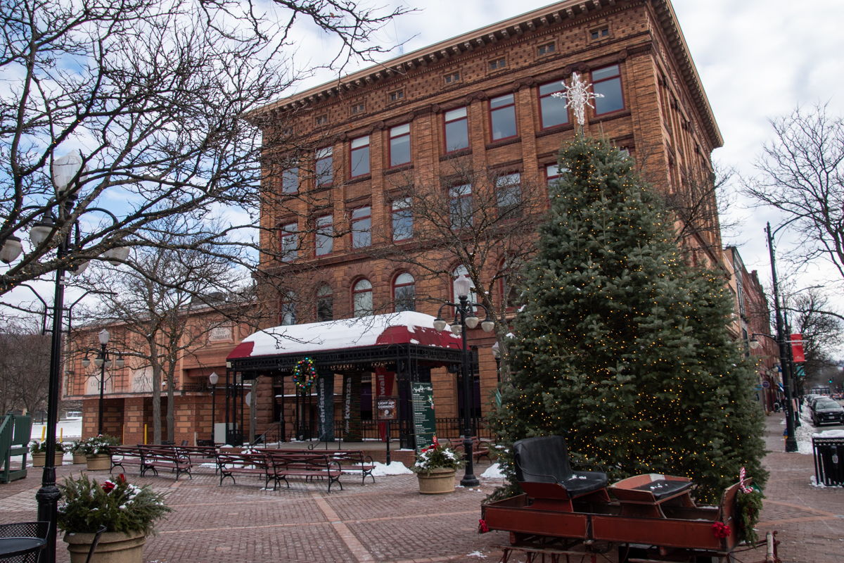A large decorated Christmas tree with warm white lights and a vintage sleigh display set along Market Street in Corning, NY, with the historic brick Baron Steuben building and snow-covered benches in the background.