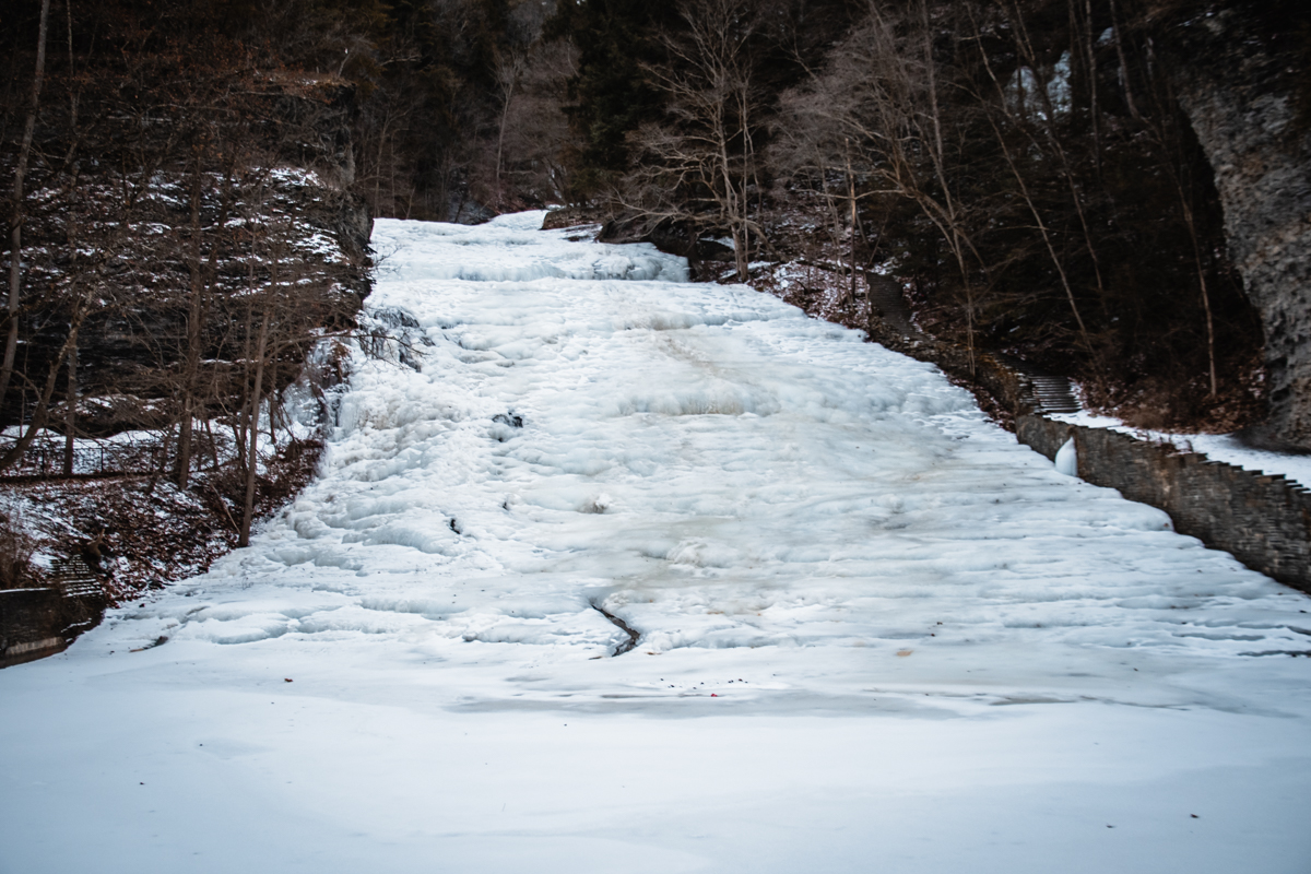 Frozen Buttermilk Falls in winter, with the lower waterfall covered in layered ice cascading down sloped rock and surrounded by snow in the Finger Lakes.