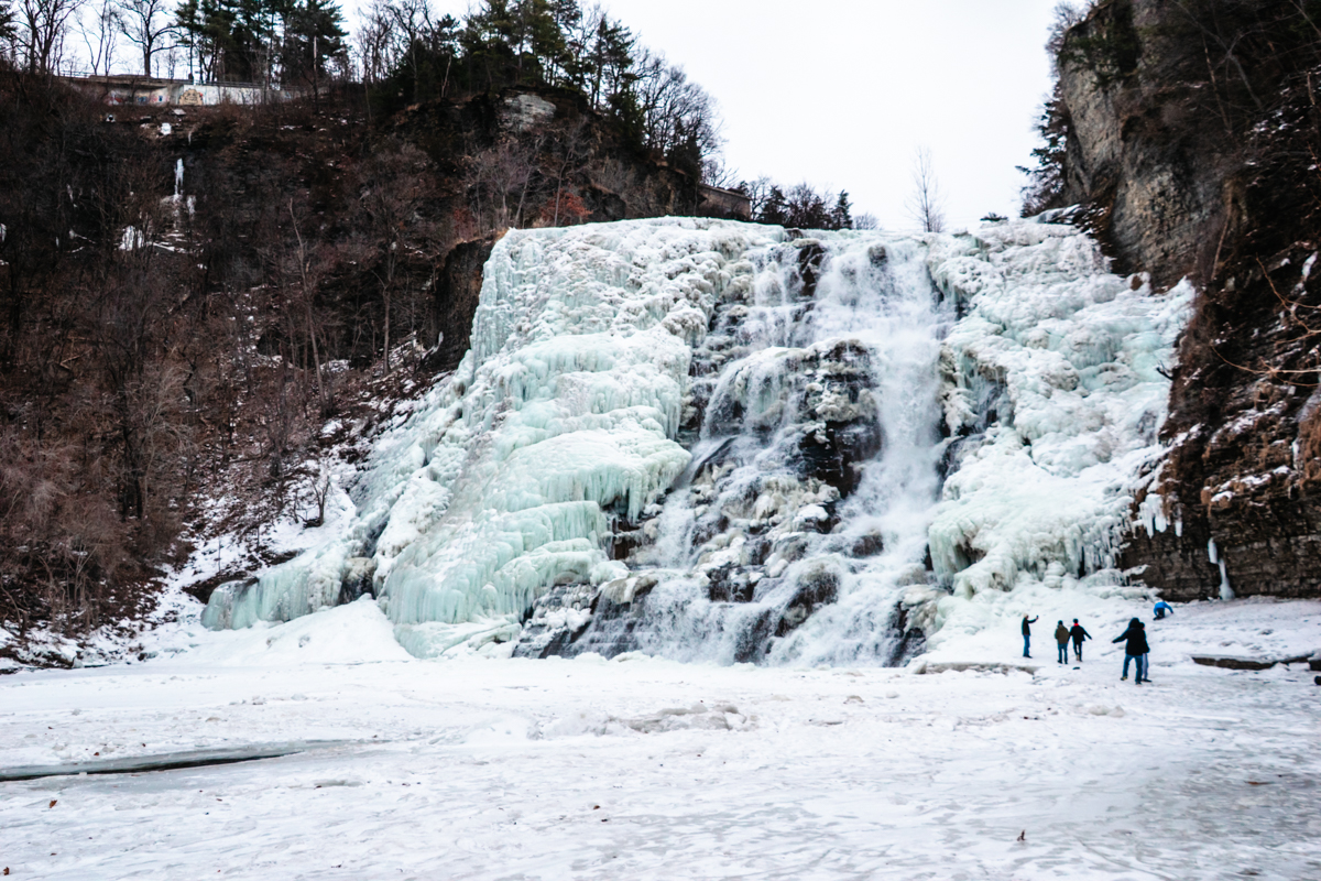 Frozen Ithaca Falls in winter, with the wide waterfall partially frozen into thick ice formations cascading over rocky cliffs.