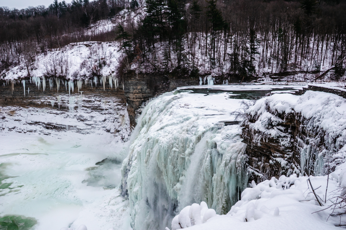 Partially frozen waterfall at Letchworth State Park in winter, showing icy cascades flowing into a snow-covered gorge below.