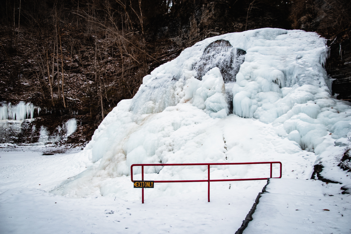 Wide view of frozen Enfield Falls in winter, with ice-covered rock formations, snowy steps, and bare trees lining the gorge.