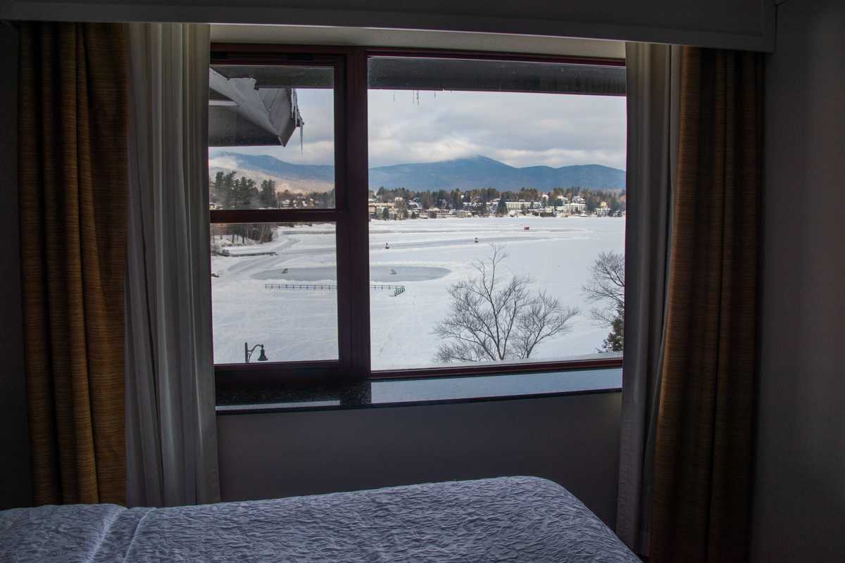 Winter view of frozen Mirror Lake in Lake Placid with snow-covered mountains seen from a hotel room window