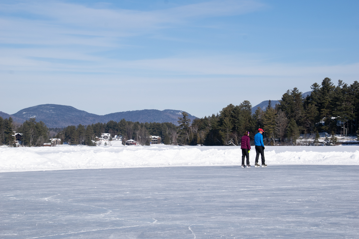Couple ice skating on frozen Mirror Lake in Lake Placid with Adirondack mountains in the background during winter