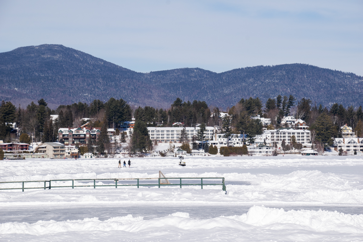 People walking across the frozen Mirror Lake in Lake Placid with snow-covered mountains and lakeside homes in winter