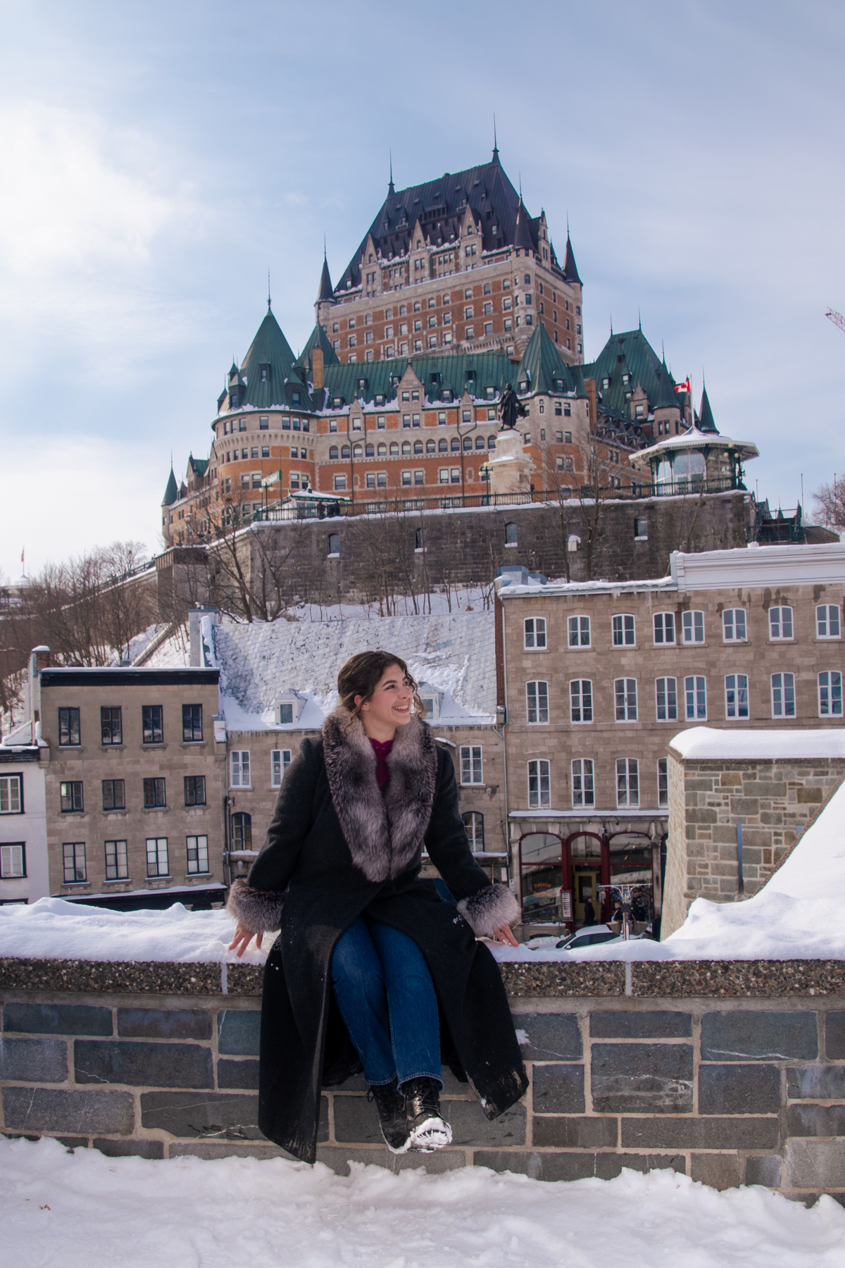 Woman sitting on snowy stone wall with Château Frontenac overlooking Old Quebec City in winter.