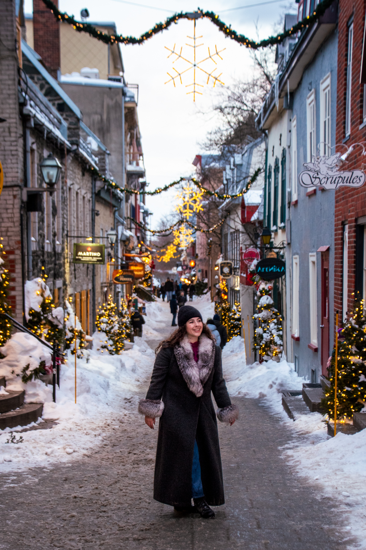 Women walking down Petit-Champlain street at dusk with glowing holiday lights and snow-lined cobblestones