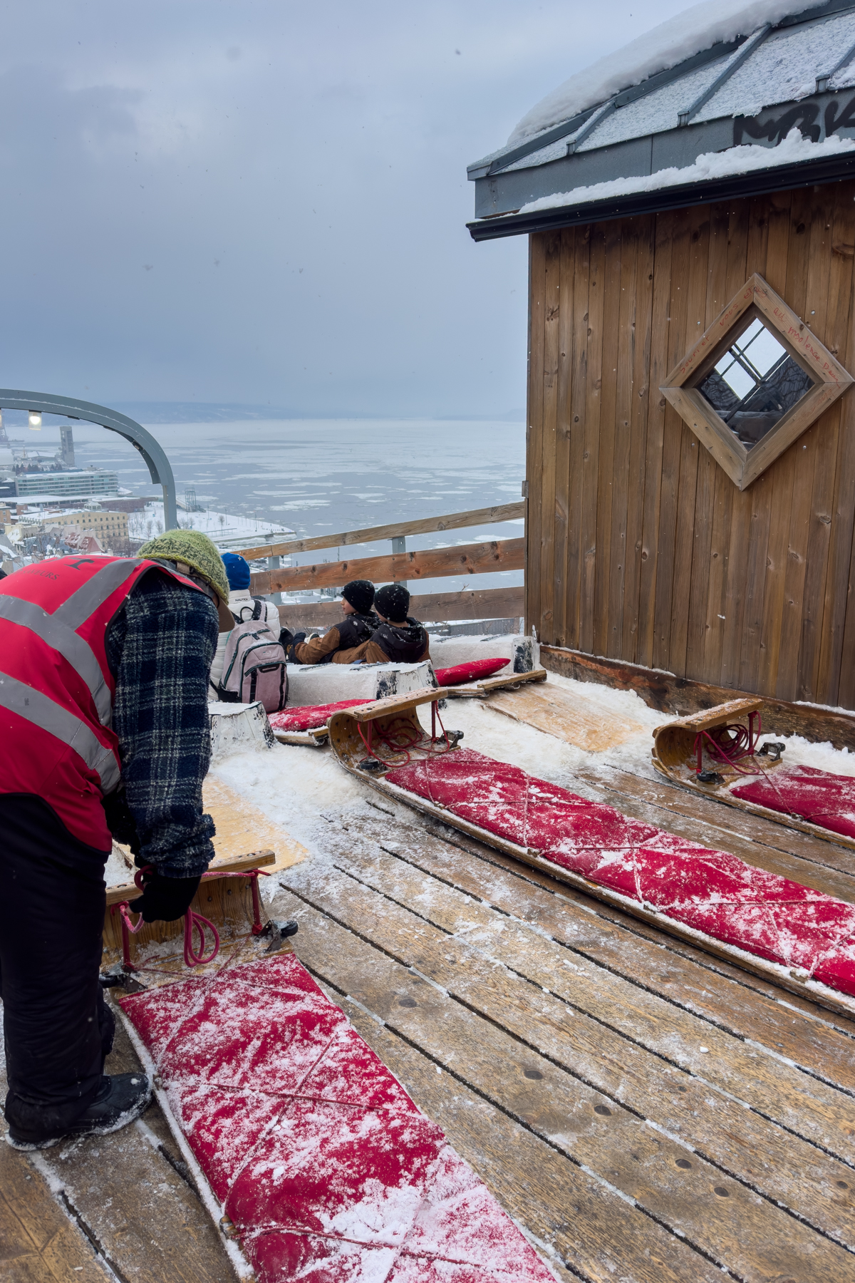 Attendant preparing wooden toboggans at the top of the Dufferin Terrace Toboggan Slide with riders seated and the St. Lawrence River below