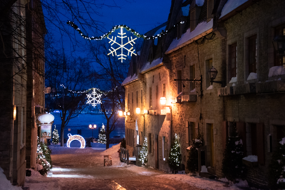 Old Quebec street at night with snowflake lights and views toward the St. Lawrence River, a romantic winter scene in Quebec City.