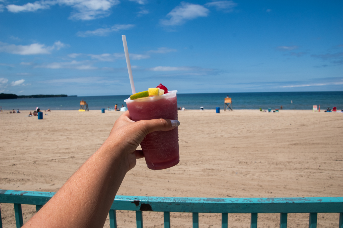 Frozen Sangria Slush from Cabana Sam's held up with views of Sunset Bay Beach and Lake Erie in the background.