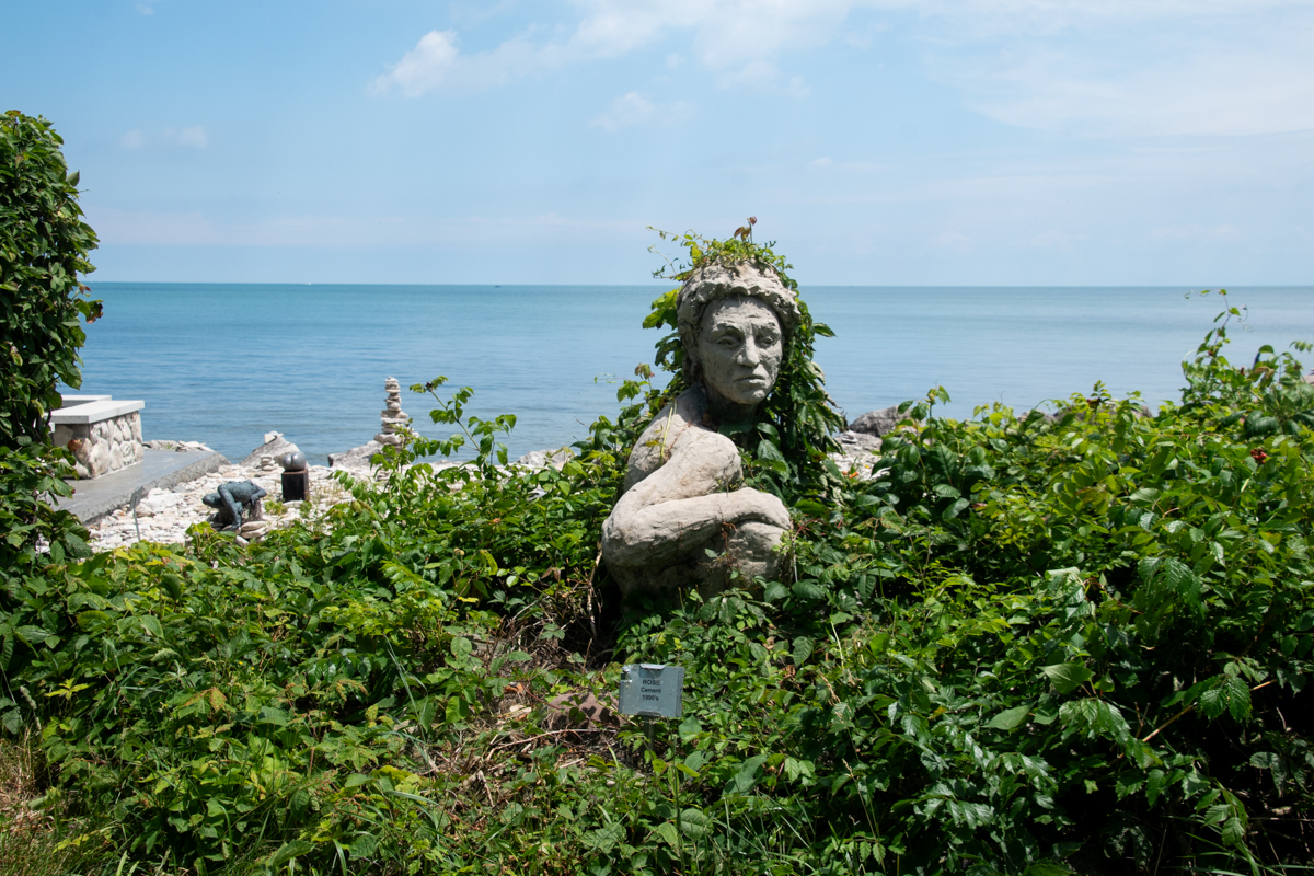Sculpture at Charles Herndon Galleries and Sculpture Garden on Kelleys Island, overlooking the rocky Lake Erie shoreline surrounded by lush greenery.