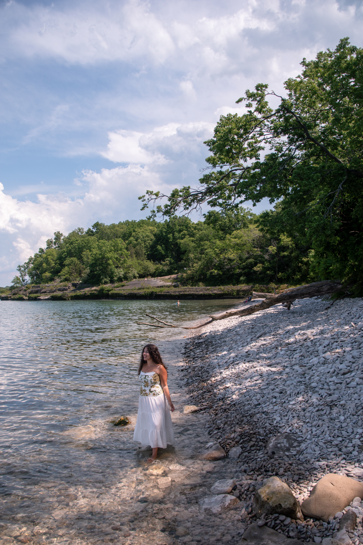 Anna Hammerschmidt of Come Join My Journey Standing along the rocky shoreline at Kelleys Island State Park, with clear Lake Erie water, limestone beach, and wooded coastline in the background.