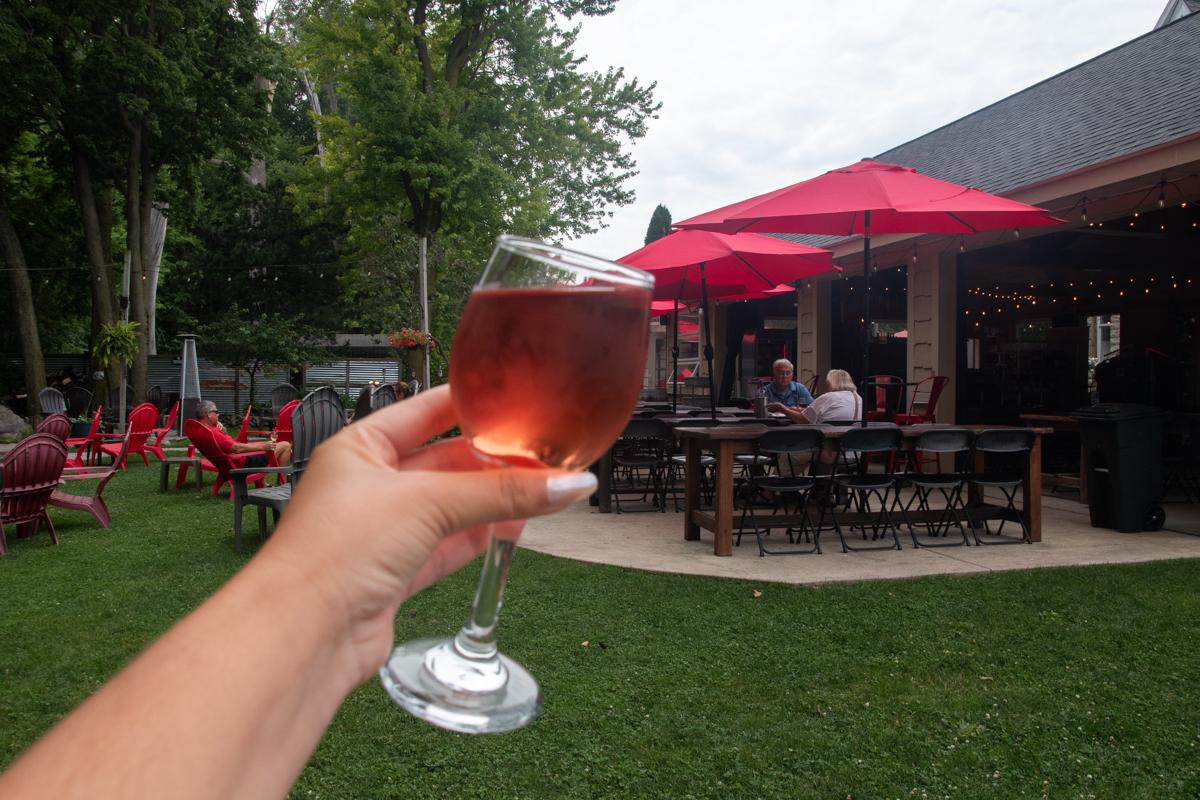 Glass of Pink Catawba wine held outside at Rocky Point Winery in Marblehead, Ohio, overlooking the winery's patio with red umbrellas and Adirondack chairs.