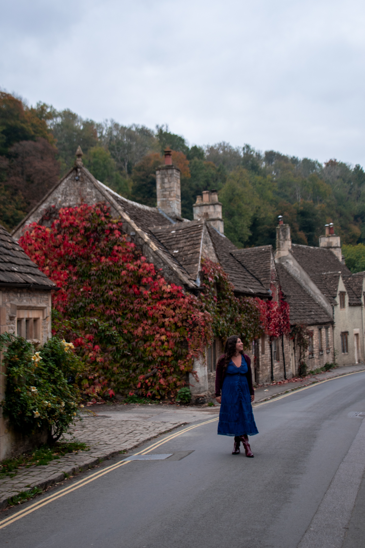 Anna Hammerschmidt of Come Join My Journey walking through streets of Castle Combe. There are stone cottages lining the road and vibrant autumn vines on the buildings.